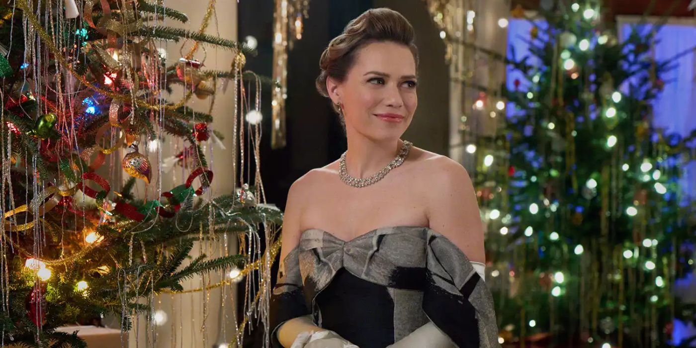 Uma foto da cintura para cima de Lucy de Bethany Joy Lutz sorrindo em um vestido de baile glamoroso e joias com enfeites iluminados cobrindo árvores de Natal atrás dela em A Biltmore Christmas da Hallmark