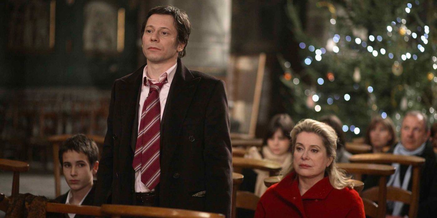 A man in a suit stands beside his mother in a small gathering of seated people with a Christmas tree in the background.