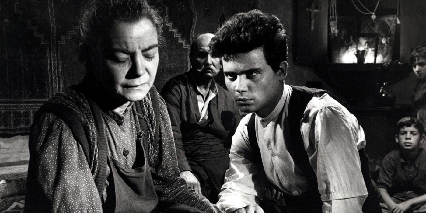 A young man sits in a crowded room, glaring at an older woman who looks down away from him.