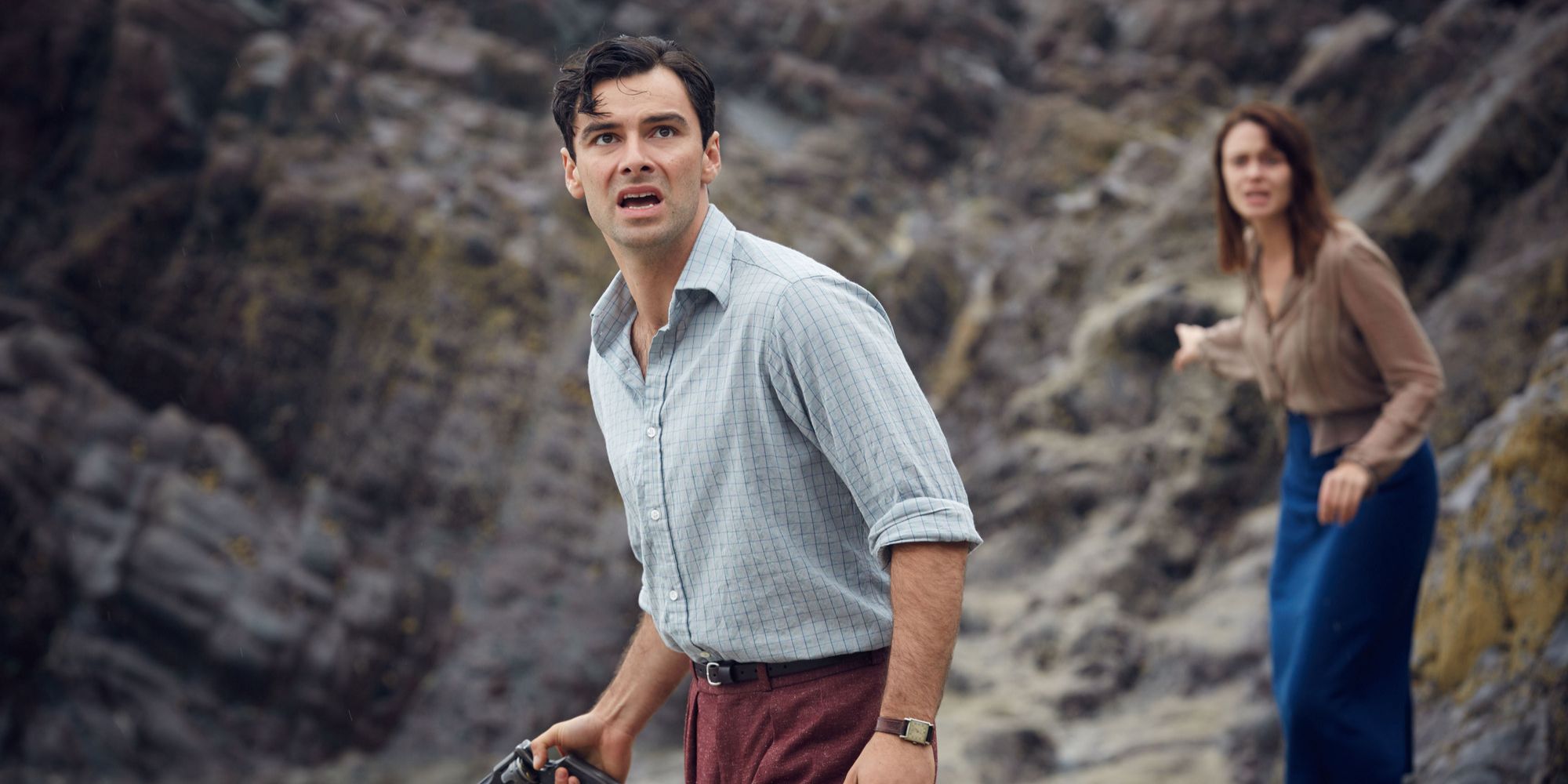 Aidan Turner's Lombard, with Maeve Dermody's Vera blurry behind him, standing amont the rockface in And Then There Were None