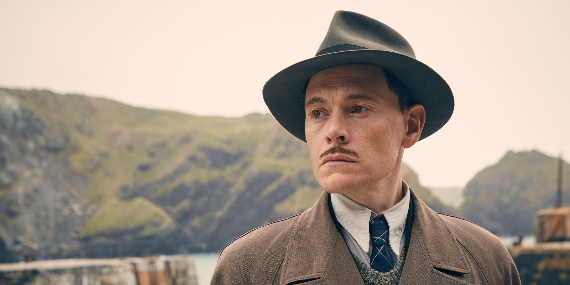 Burn Gorman as William Blore staring to the left wearing a hat and standing outside with the island mountains in the distance in And Then There Were None