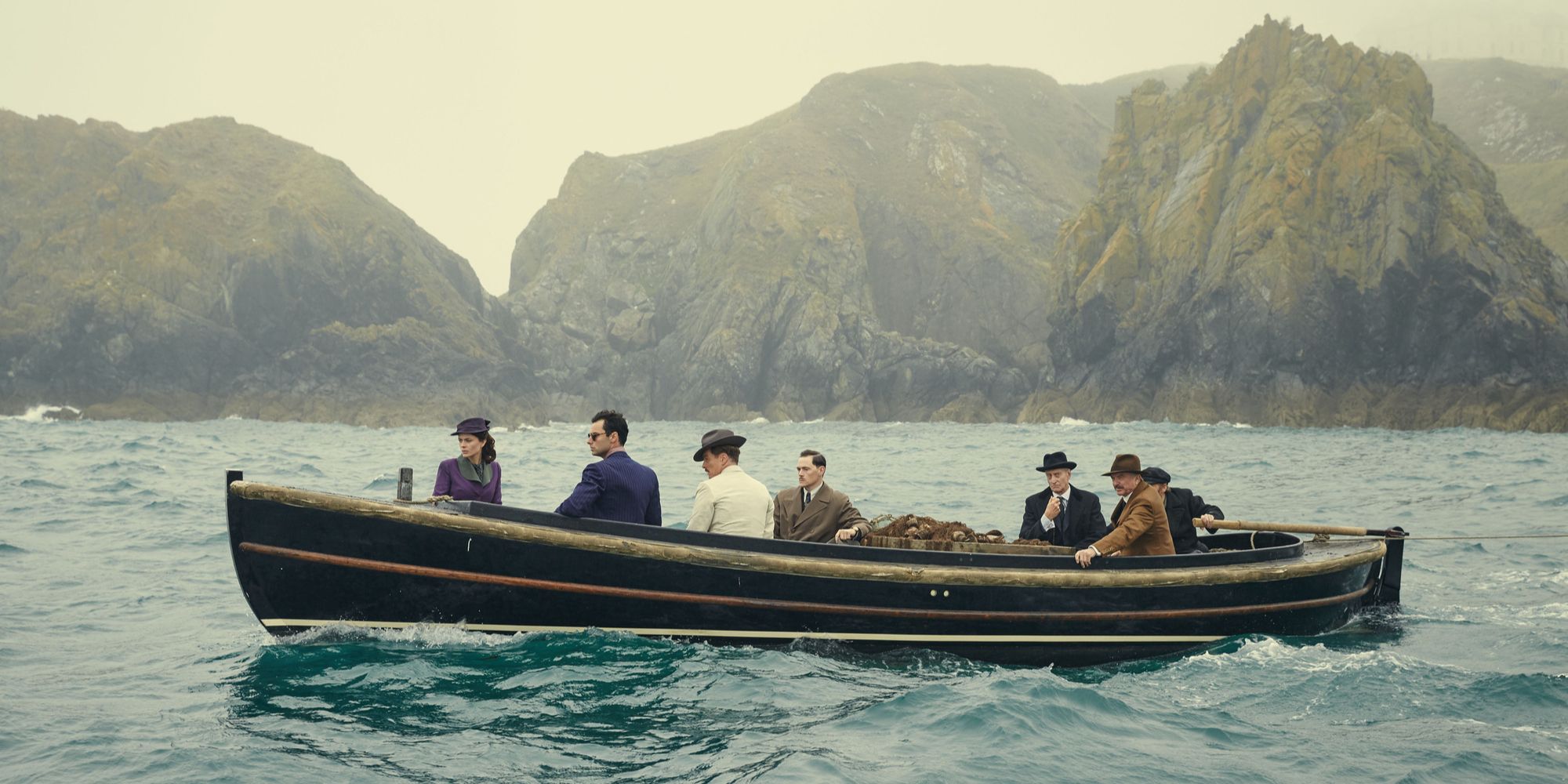 A wide shot of the cast of And Then There Were None as they sail a boat across choppy seas with island hills behind them