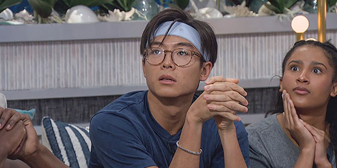 Derek Xiao from Big Brother sitting on a couch with a blue headband looking up at the screen.