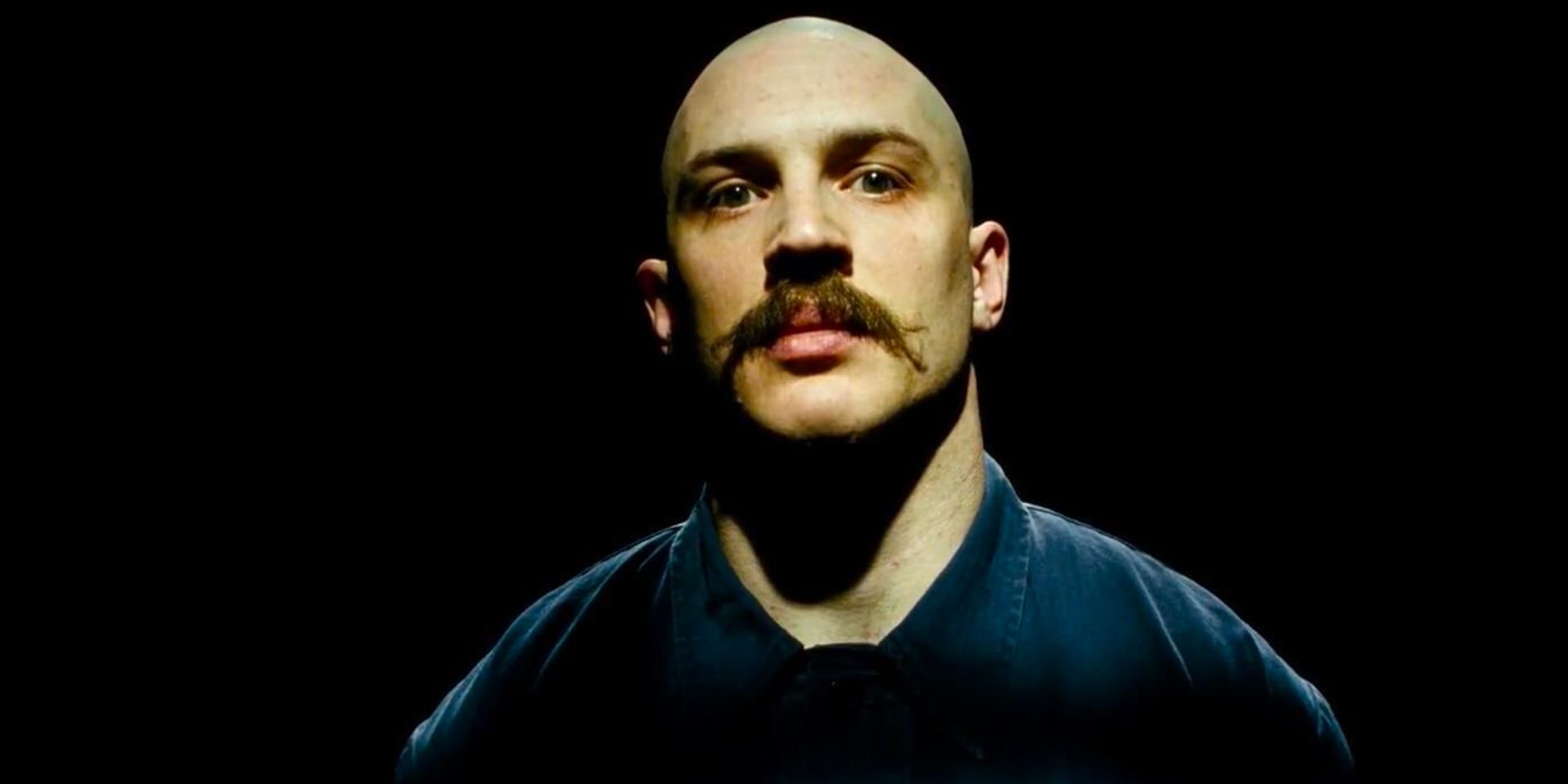 Michael Peterson aka Charles Bronson (Tom Hardy) stands in a spotlight in an otherwise blackened room looking down the barrel of the camera.