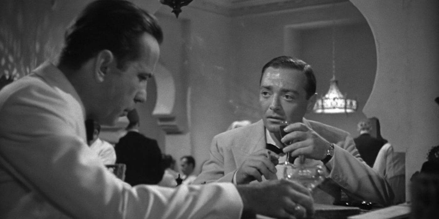 Two men meet in a nightclub with one sipping on his drink and smoking while the other looks down despondently.