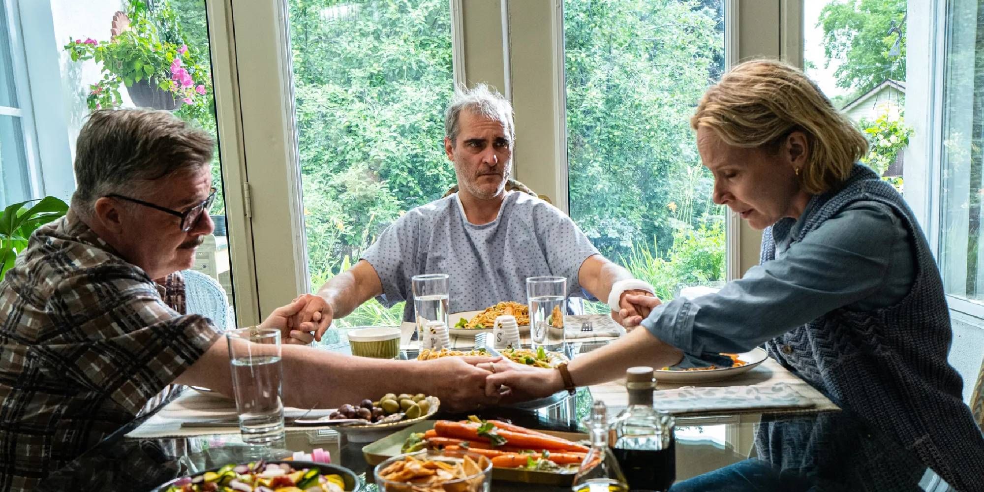 Joaquin Phoenix, Nathan Lane, and Amy Ryan holding hands at the table in Beau is Afraid.
