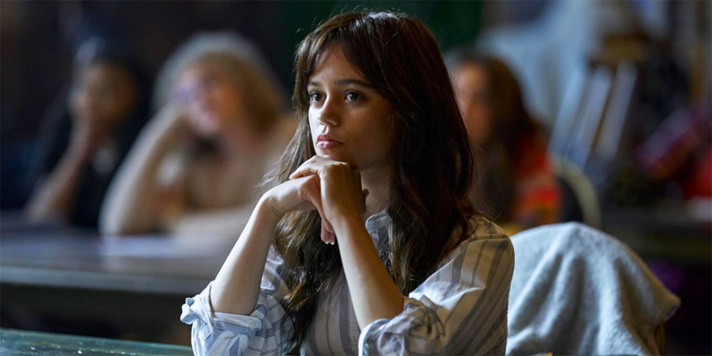 Jenna Ortega sitting at a desk in a classroom, her hands under her chin