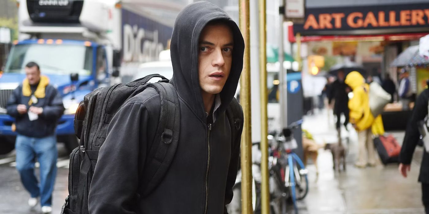 Rami Malek as Elliot Alderson wearing a hoodie and looking to his right on a crowded street in Mr. Robot.