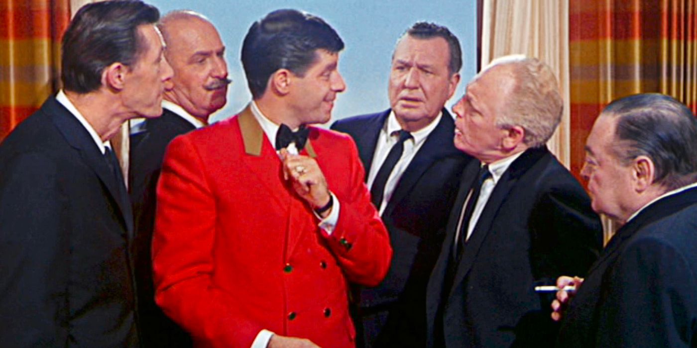 A young, joyous bellhop in a bright red jacket stands surrounded by comedy managers in a hotel room.