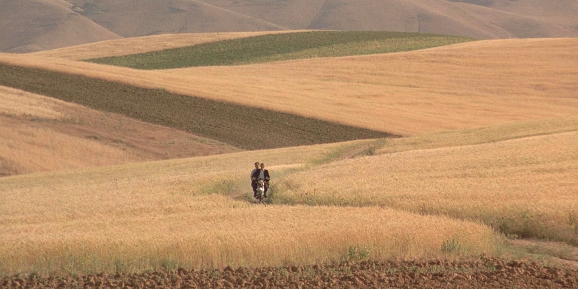 Two figures ride a motorcycle across a wheat field in The Wind Will Carry Us