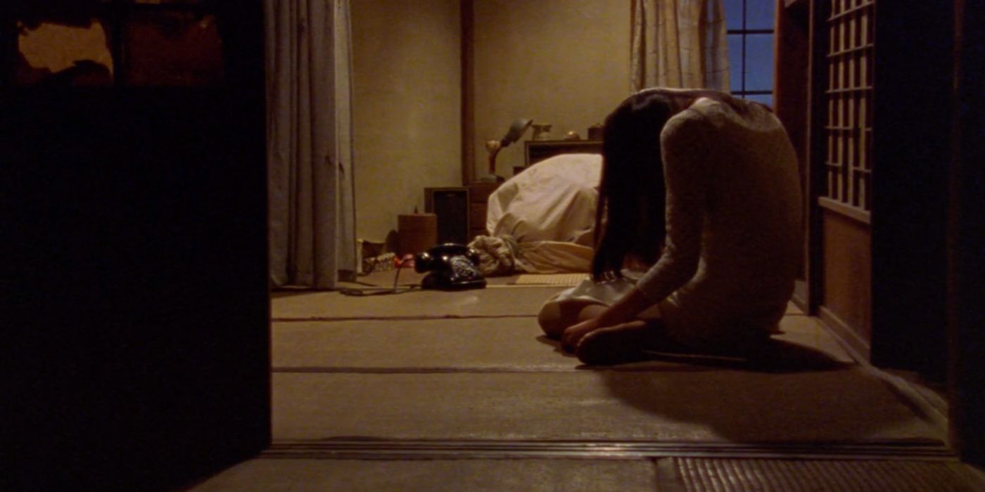 A girl sits hunched on the ground in a room with a phone and a bag on the ground in front of her.
