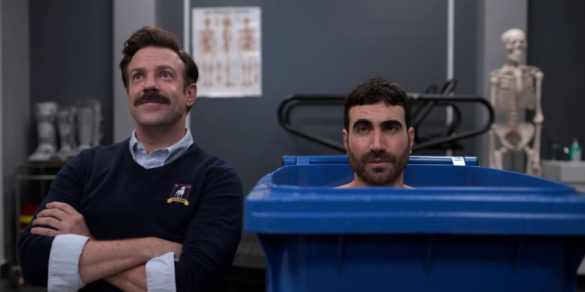 Jason Sudeikis is sitting on a chair and smiling next to a grumpy Brett Goldstein having an ice bath inside a blue trash can in Ted Lasso