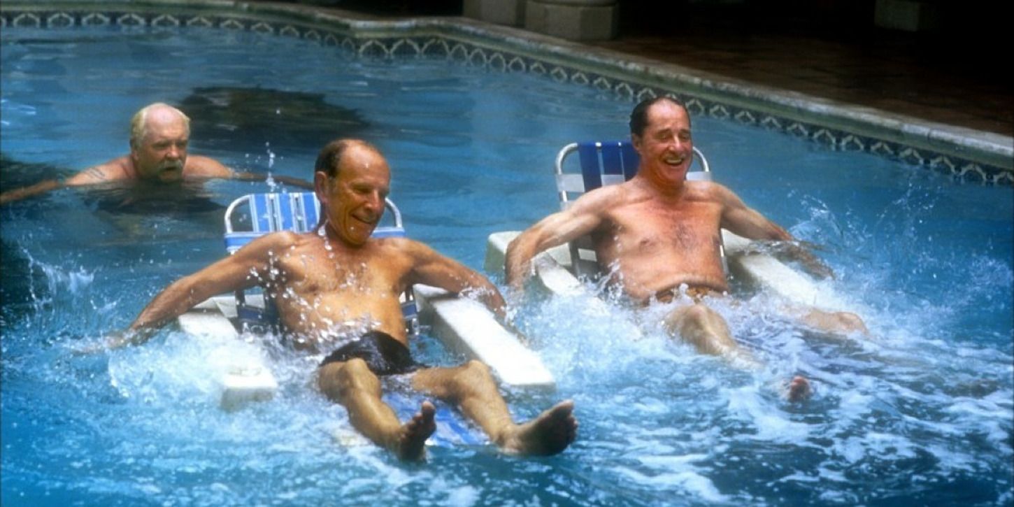 two men on floating chairs in a pool, another man swimming behind them