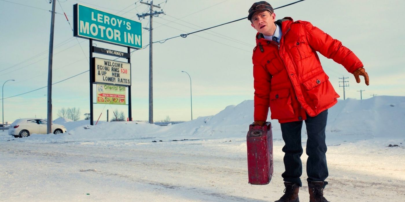 A man in a bright orange jacket stands in the snow with a gas can by a highway motor inn.