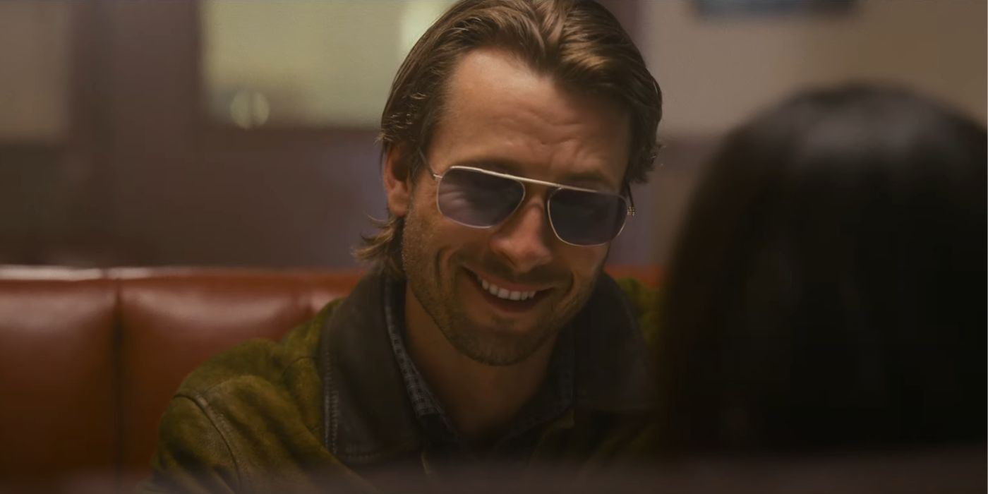 Glen Powell as Gary Johnson, sitting in a restaurant and smiling while wearing Hit Man sunglasses