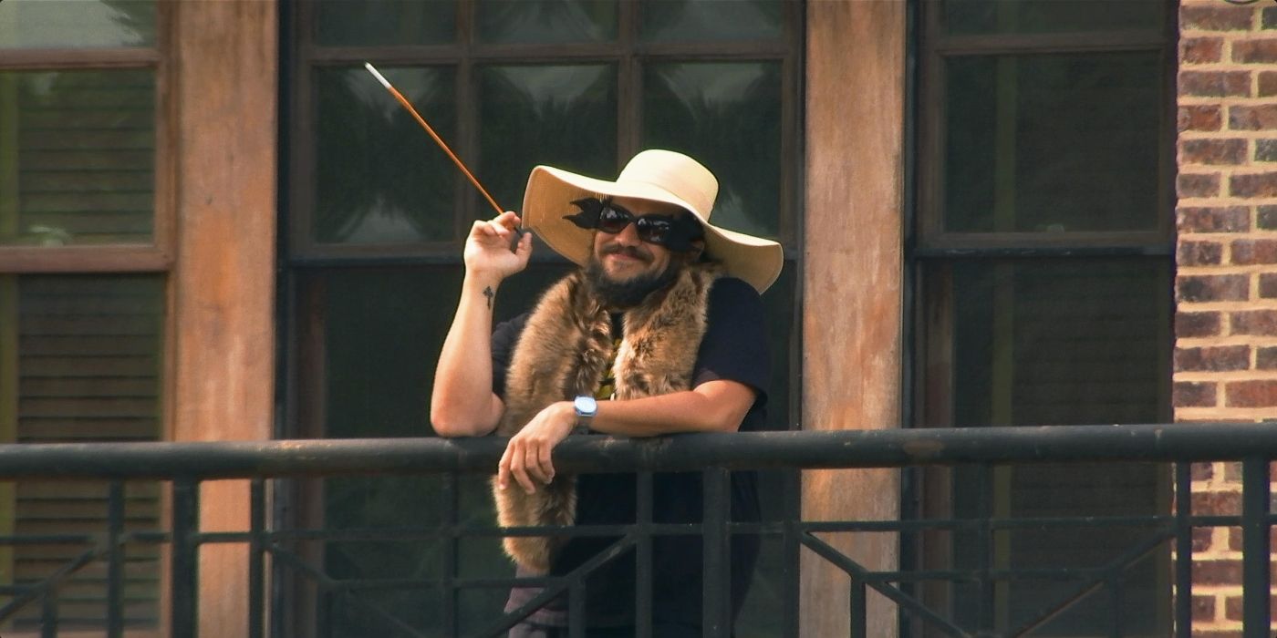 Sal Vulcano on a balcony wearing a sunhat and using a long cigarette holder like Cruella Deville