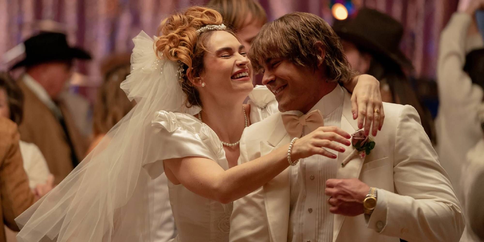Lily James as Pam Adkisson in a wedding gown holding Zac Efron as Kevin Von Erich and laughing in The Iron Claw