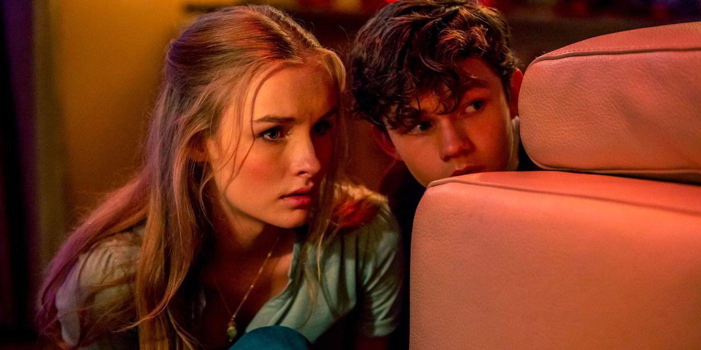 A young boy and his teenage babysitter hide behind a pink sofa, peering over it. 