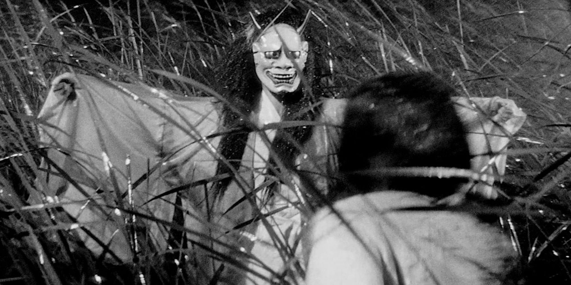 Man in a devil mask standing in front of a girl in a field in Onibaba