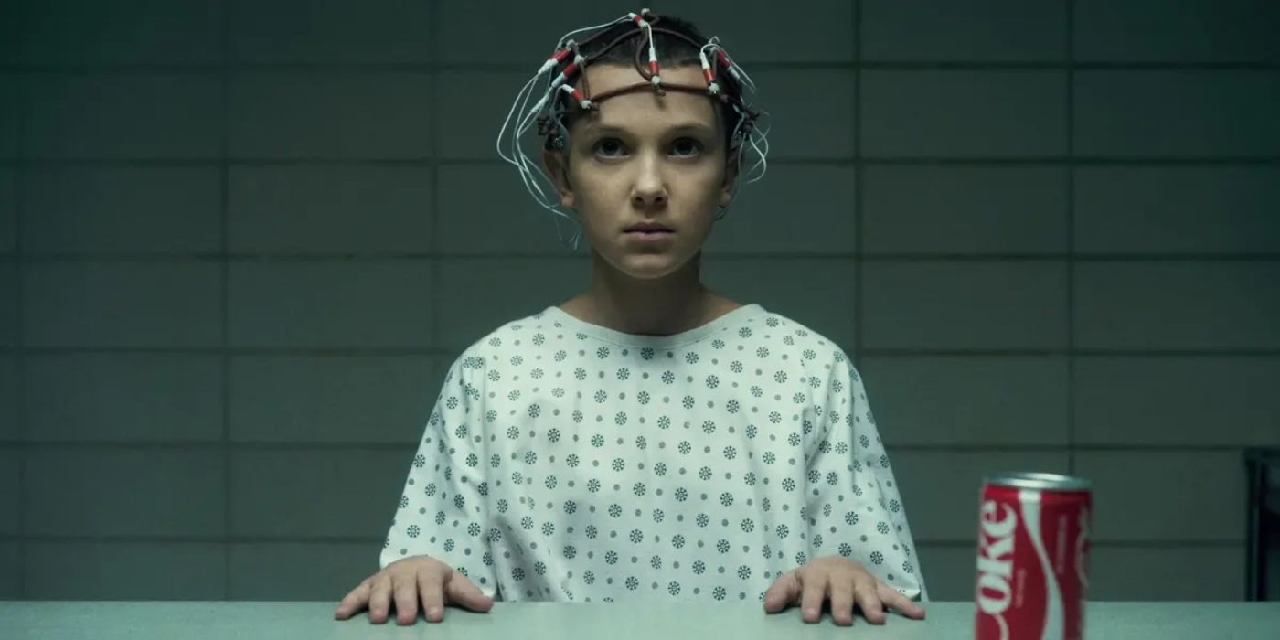 Eleven wearing electrodes on her head, sitting at a table with a coke can in Stranger Things.