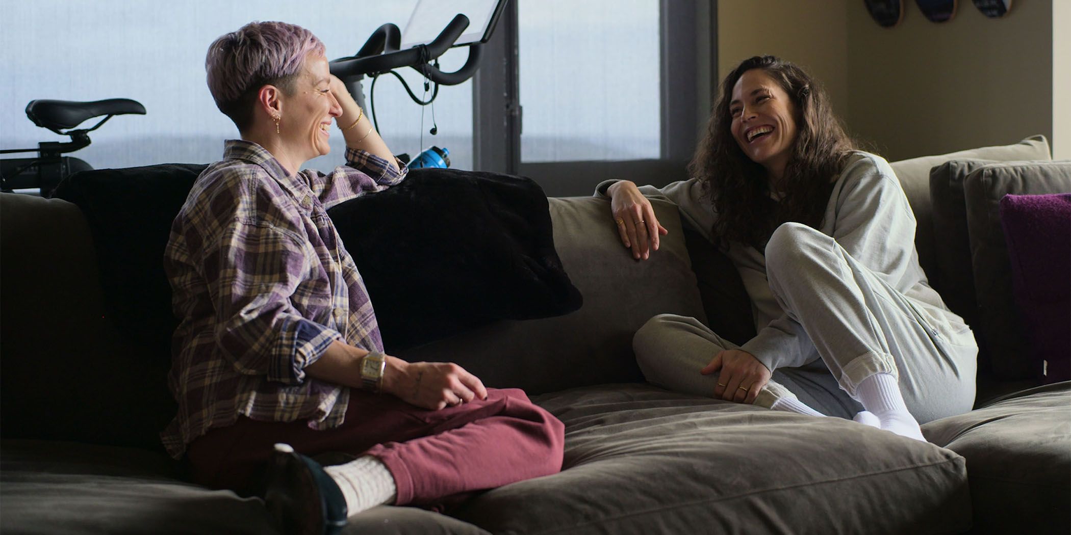 Megan Rapinoe (left) and Sue Bird (right) laughing while sitting on a couch in Sue Bird: In the Clutch for Sundance 2024. 