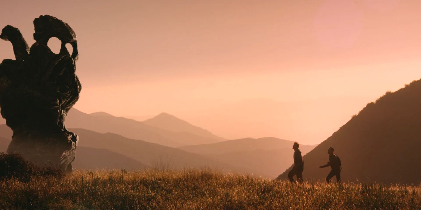 Two people walk across a grass landscape amid the mountains, nearing a mysterious large figure.