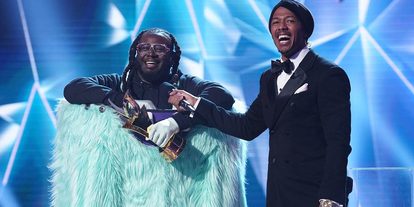 Nick Cannon on stage laughing, holding a mic up to T-Pain in costume on The Masked Singer.