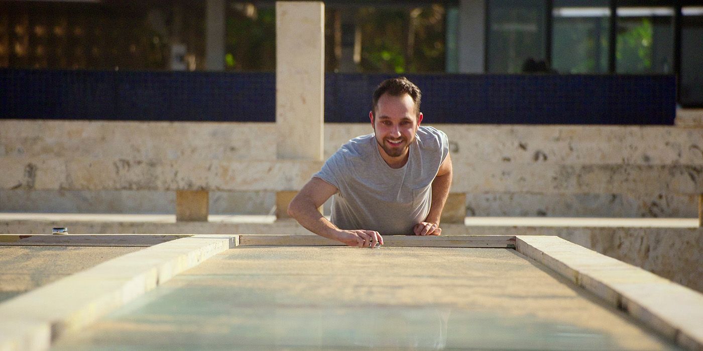 Gaspare from The Trust: A Game of Greed at the end of a shuffleboard table, about to hit the disc and smiling.