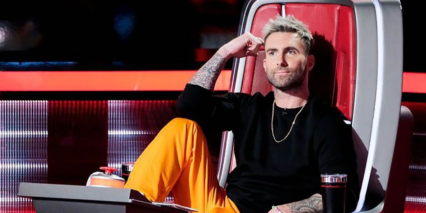 Adam Levine sitting in his chair on The Voice, leg up and arm to his head smiling.