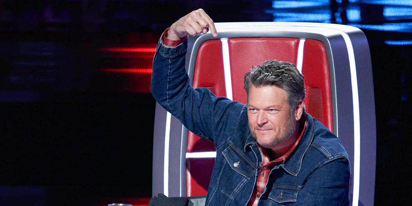 Blake Shelton sitting in his chair on The Voice, pointing his finger to his head.