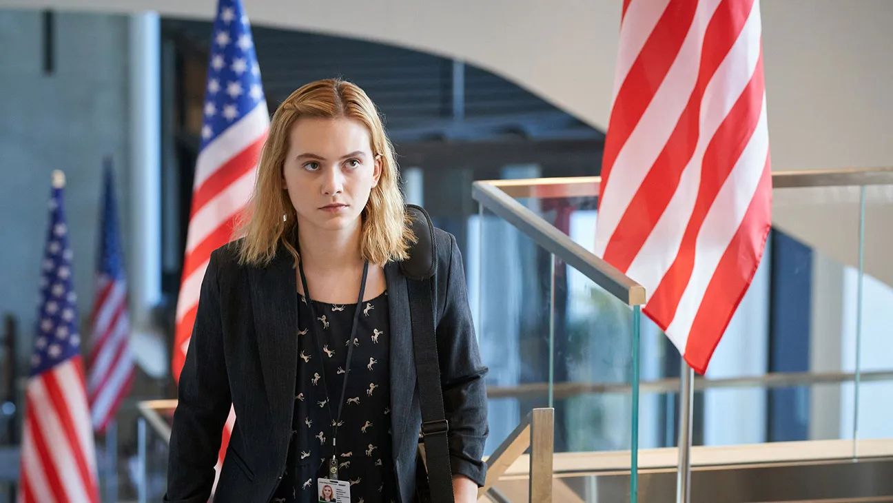 Emilia Jones wearing an employee badge walking up the stairs as Reality Winner with American flags in the background in Winner