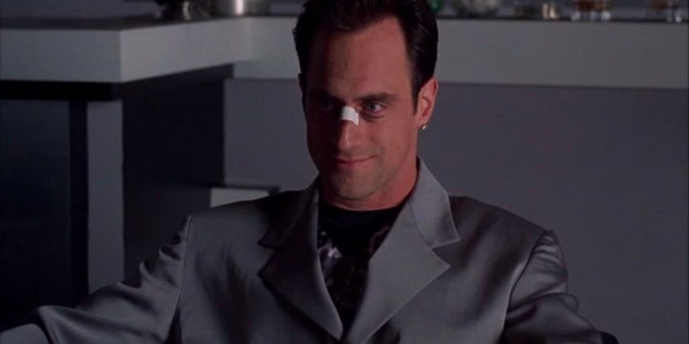 A bruised thug, Johnnie Marzzone (Christopher Meloni) sits smiling while wearing a grey suit.