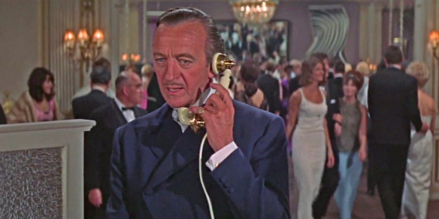 James Bond (David Niven) speaks on the phone in a crowded lobby area.