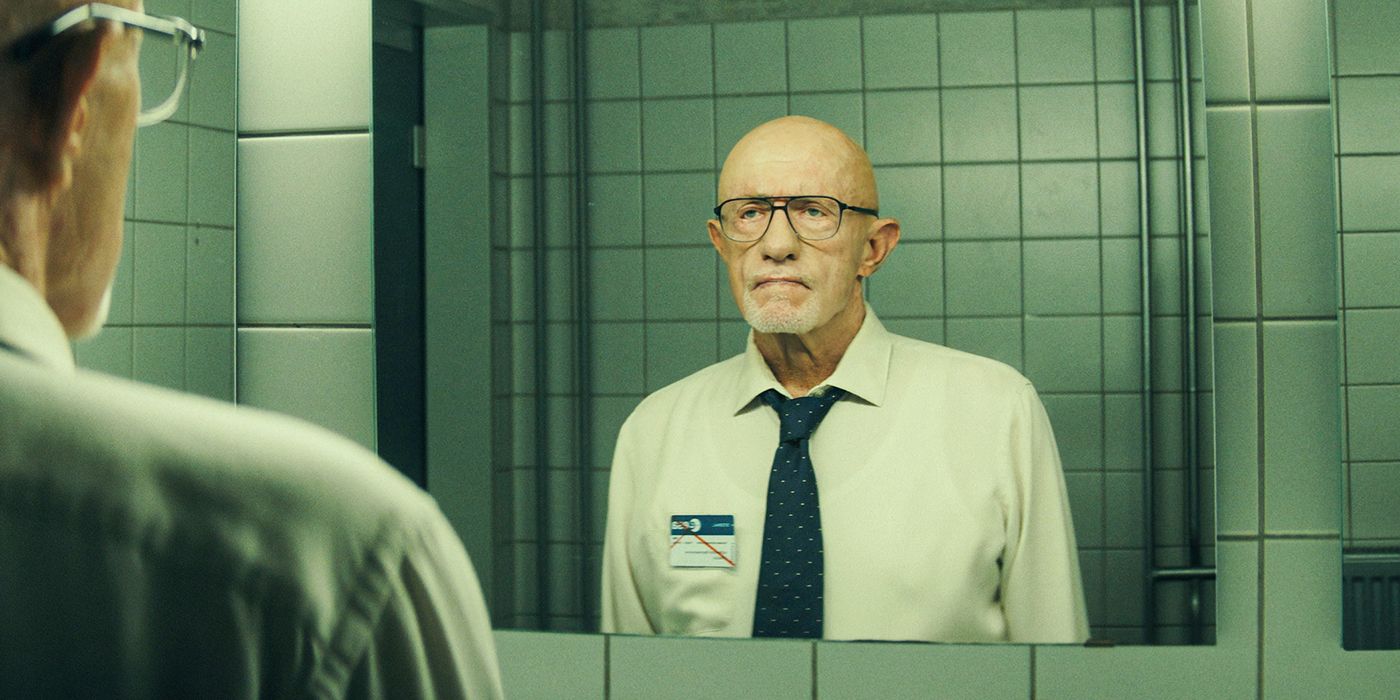 Jonathan Banks as Henry Caldera looking at a mirror while wearing glasses, a tie, and a name-tag with a grim expression in Apple TV+'s Constellation. 