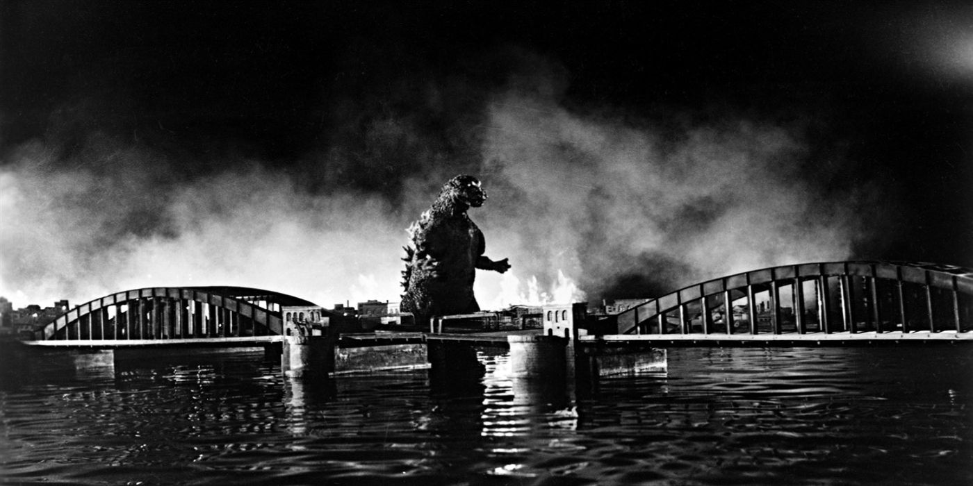 Godzilla crosses a river, with a bridge in the forefront and smoke from his destruction behind in 'Gojira' (Godzilla) 1954