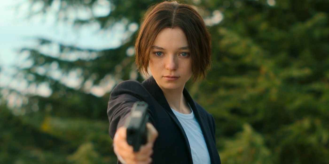 Hanna (Esme Creed-Miles), a young woman, aims a gun down at someone in a serene, tree-filled setting.