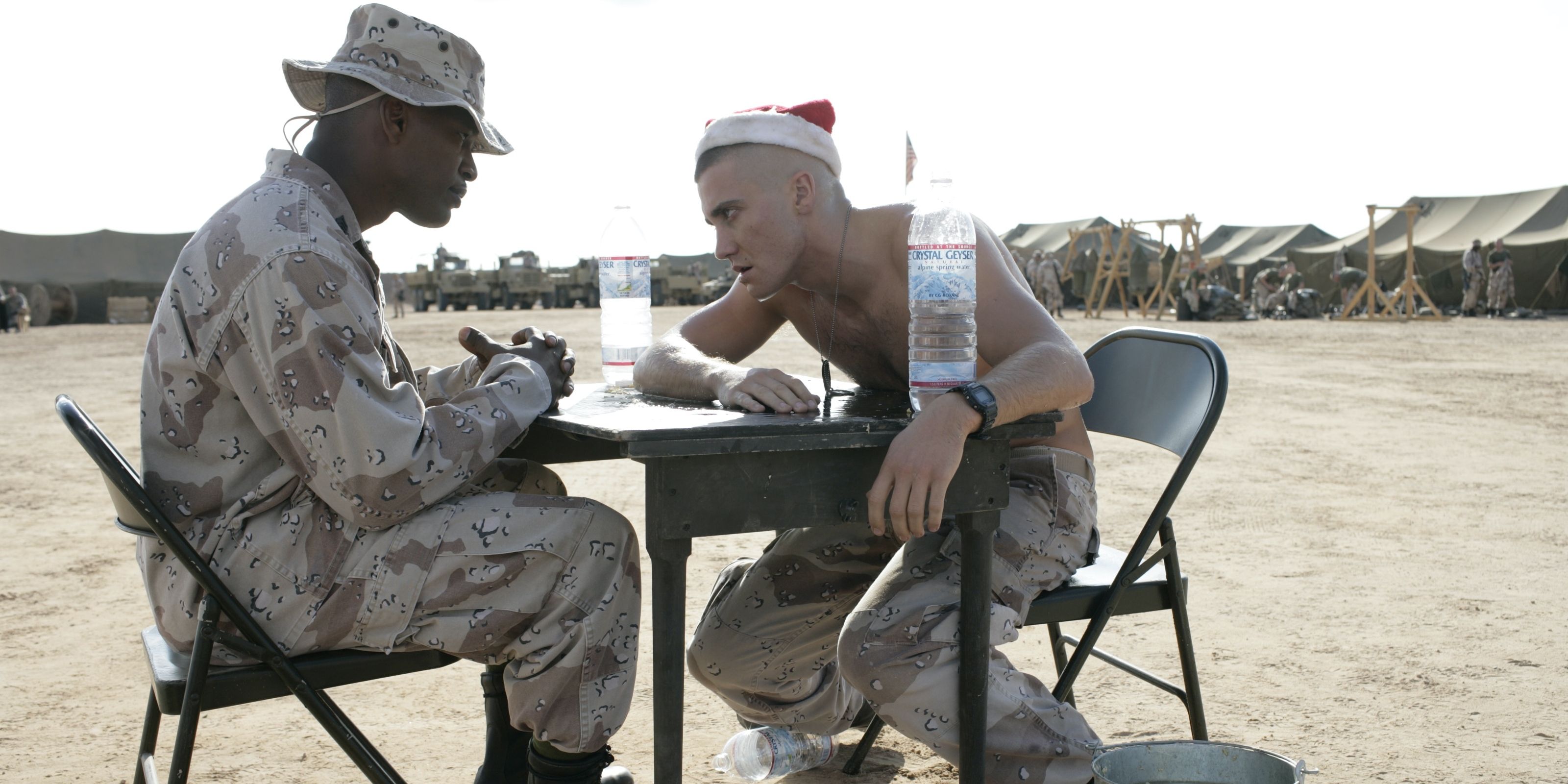 Two soldiers sitting on a table in the desert, one in full camo, the other shirtless wearing a Santa hat