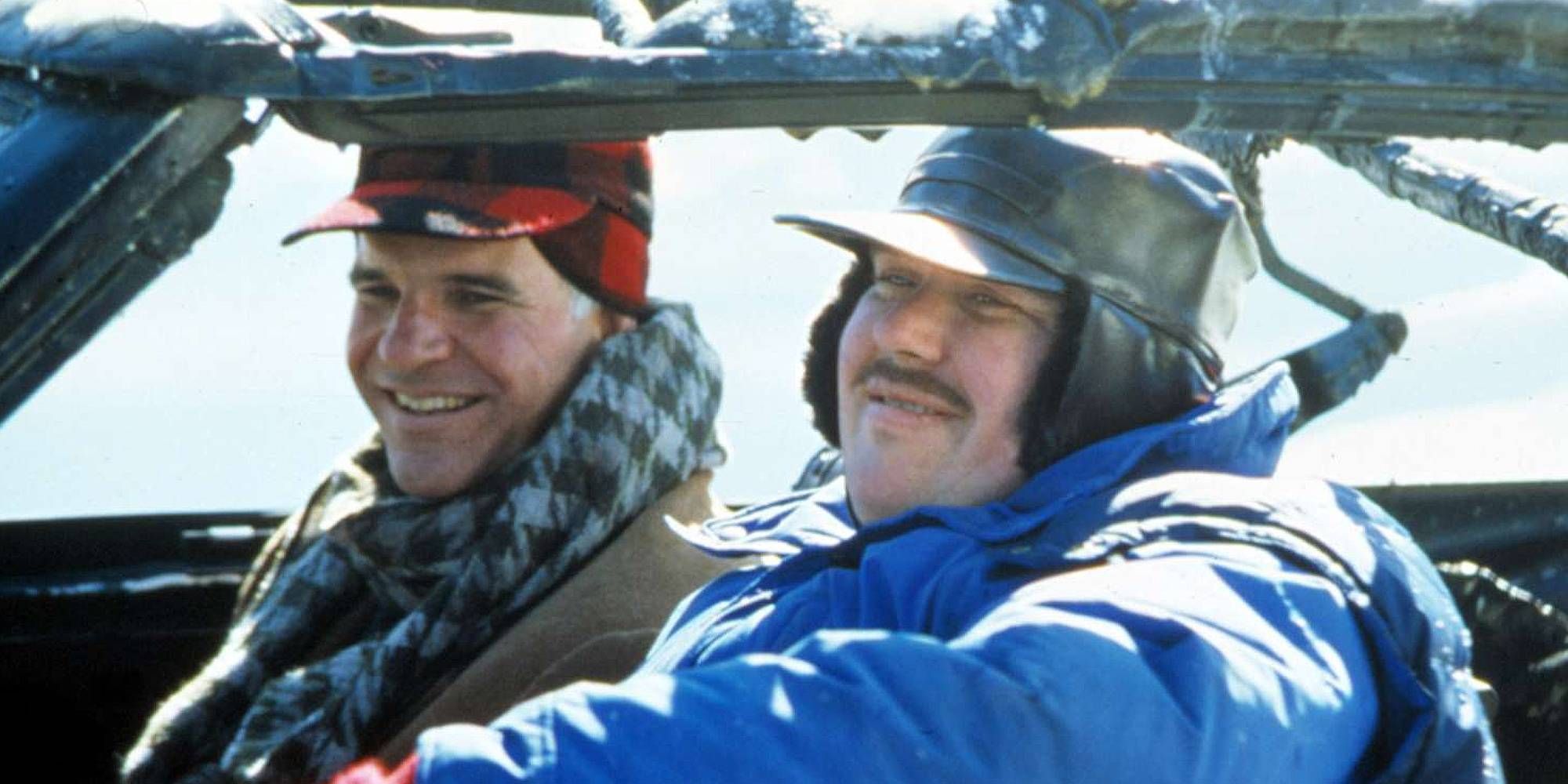 John Candy and Steve Martin in Planes, Trains, and Automobiles are smiling sitting next to each other inside a car.