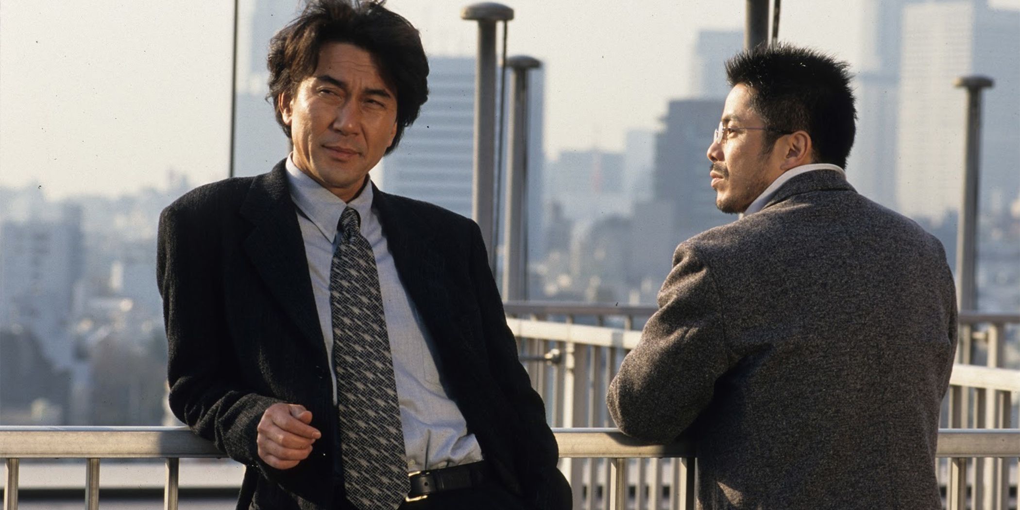 Koji Yakusho as Kenichi Takabe in a suit leaning against a guardrail with a city skyline in the background in Cure.