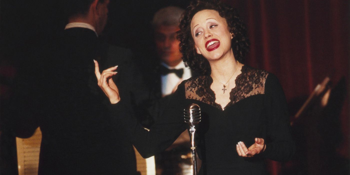 Edith Piaf (Marion Cotillard) sings into a microphone on stage while wearing a black dress and a cross necklace.