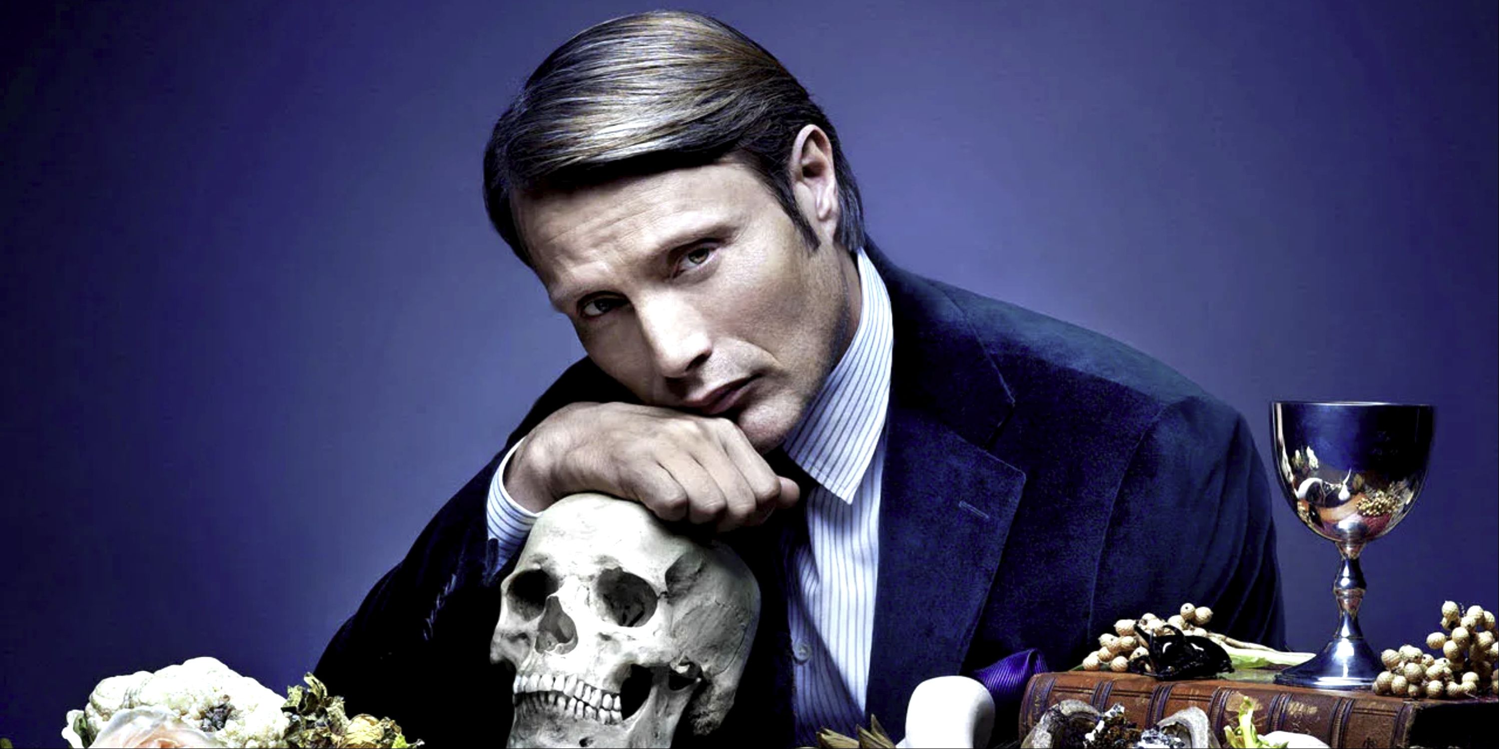 Mads Mikkelsen as Hannibal Lecter leans on a skull at the dinner table.