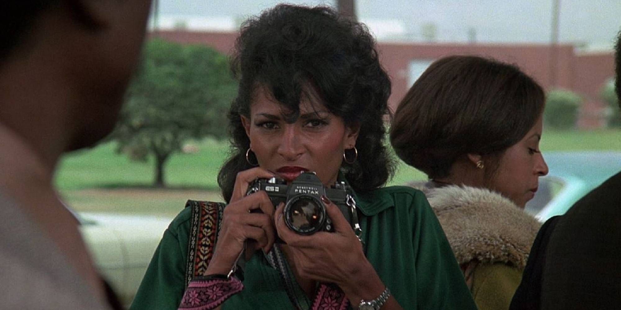 Pam Grier in Friday Foster holding a camera while looking at someone off-camera.