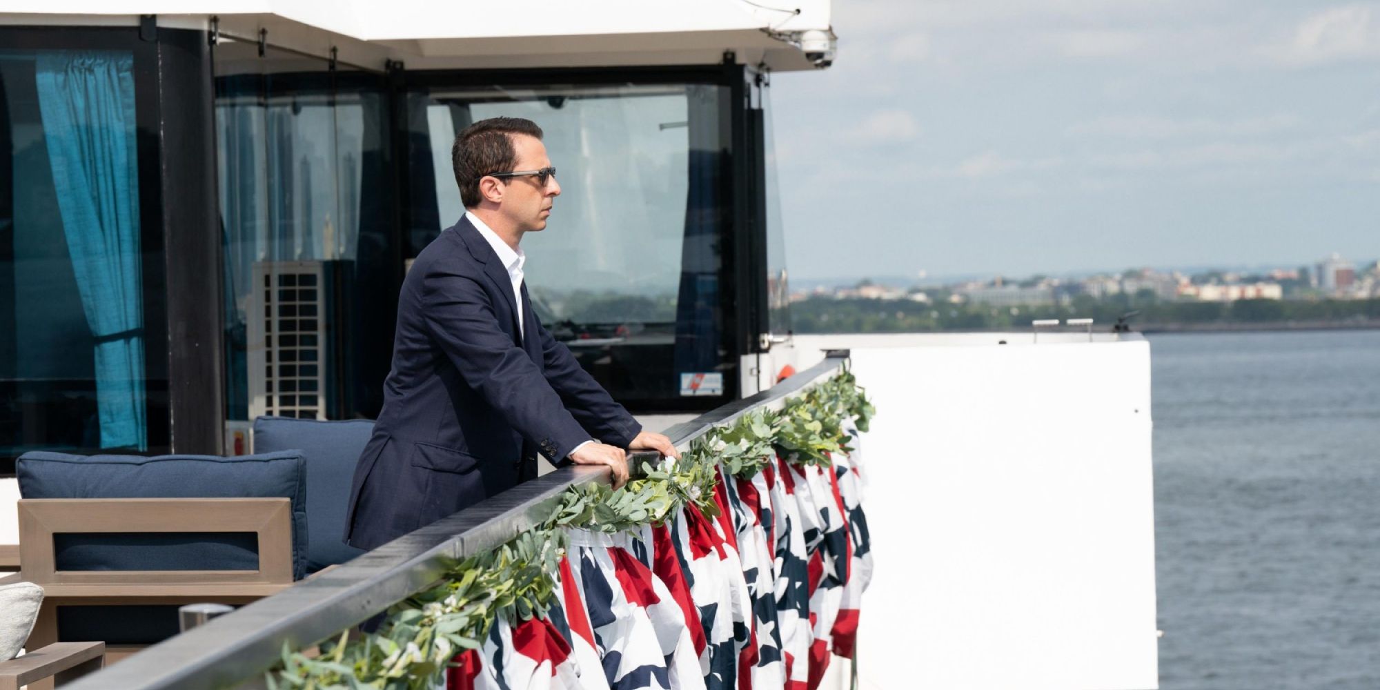 A man stands on a decorated boat on the water, looking over the edge into the distance in Succession.
