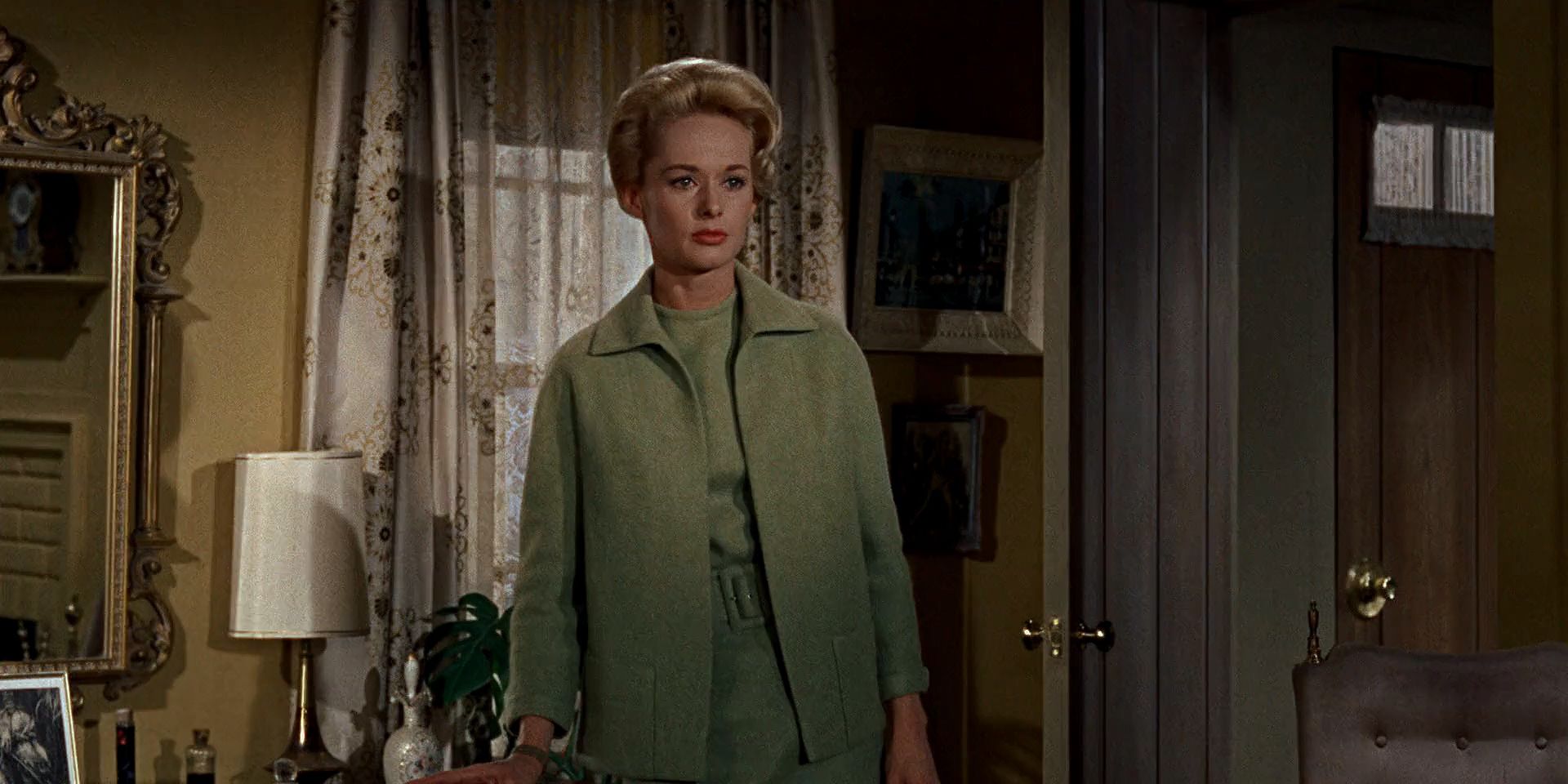 Melanie Daniels, played by Tippi Hedren, standing in a room looking ahead and wearing a matching light green jacket, shirt, skirt, and belt in The Birds