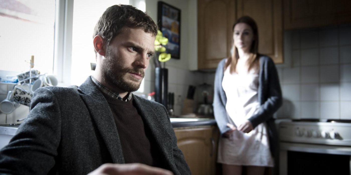 Jamie Dornan as Paul Spector sitting down in a kitchen with a woman standing behind him to his right in The Fall