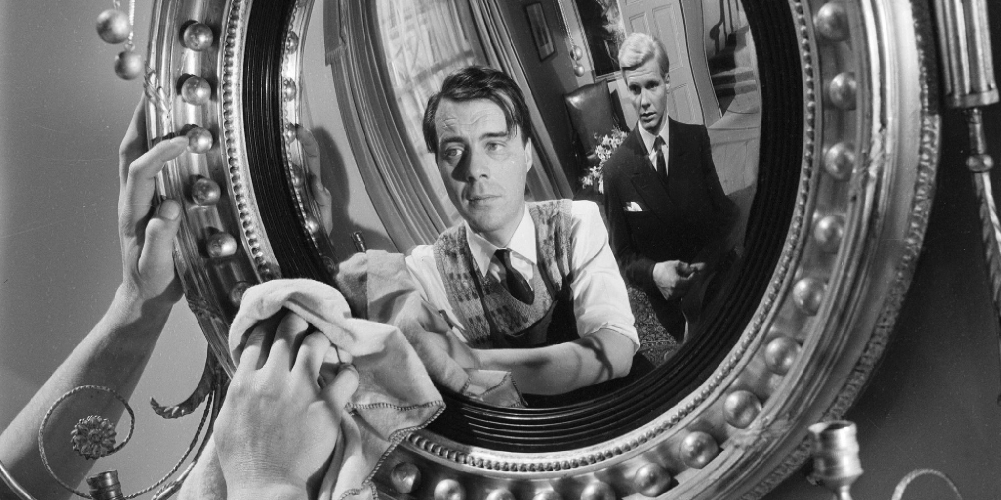 A man cleaning a mirror while another man stands behind in The Servant - 1963