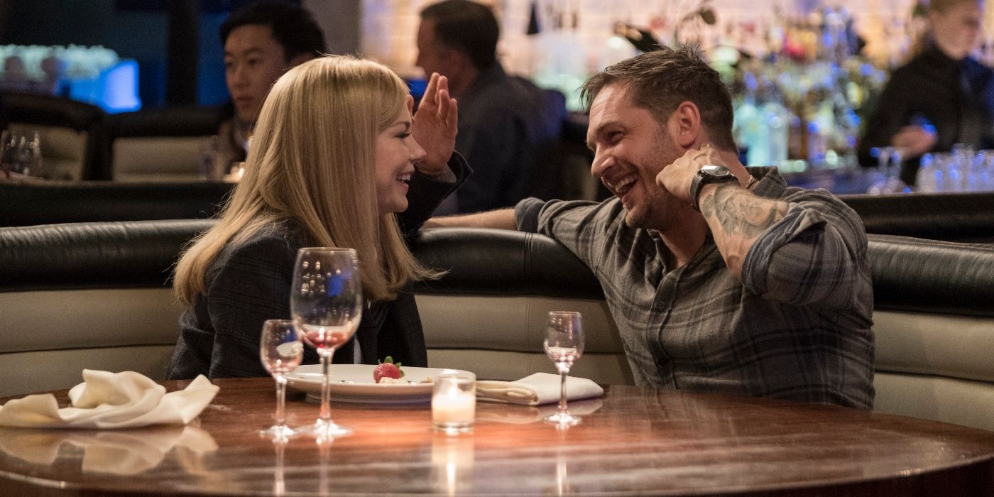 Tom Hardy and Michelle Williams as Eddie Brock and Anne Weying, sitting at a dinner and laughing in Venom.