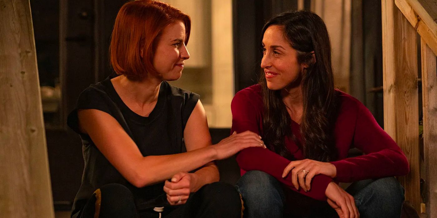 Two women sit on the front porch at night, smiling at each other as they share a friendly, supportive moment.