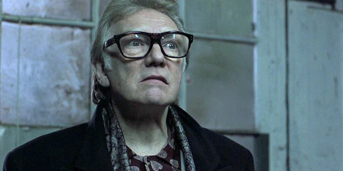 Brick Top (Alan Ford) stands in a grim industrial building wearing large glasses and grimacing in enraged disgust.