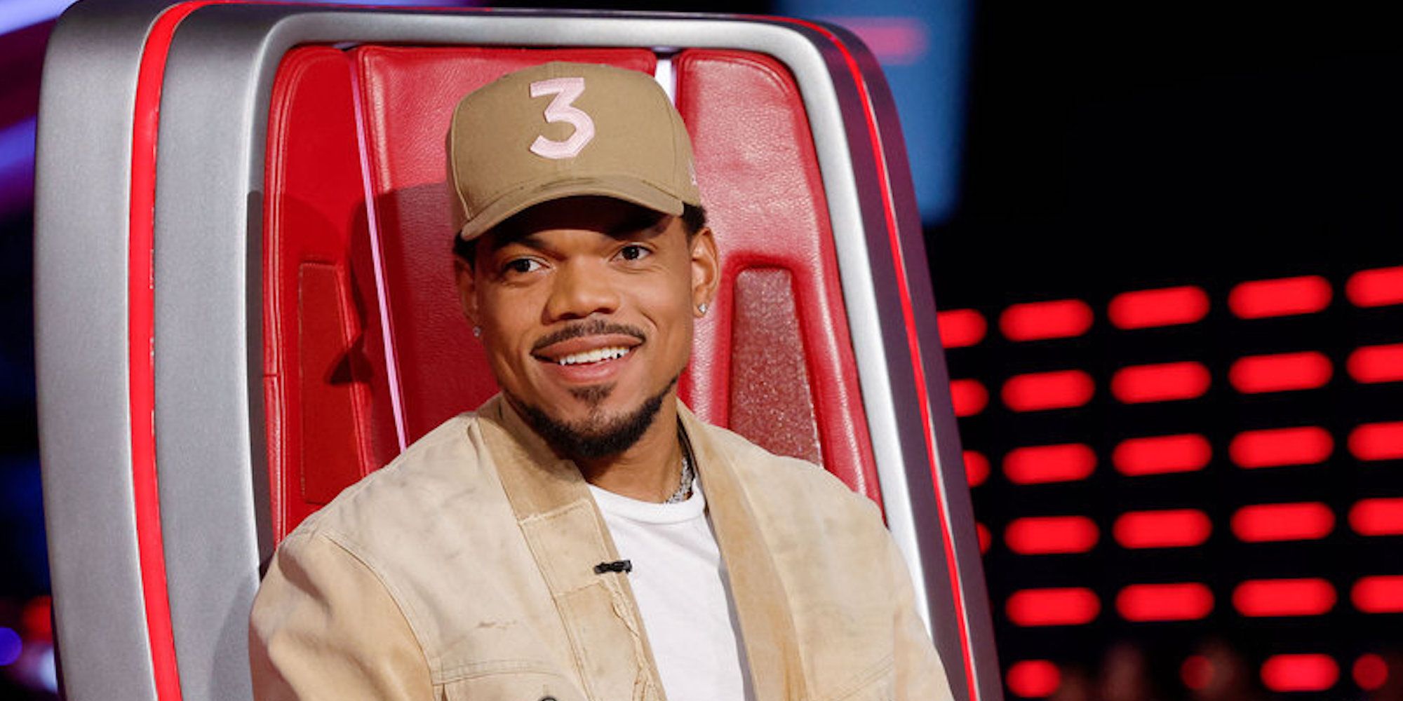 Chance the Rapper looks toward the camera smiling while seated in his chair on 'The Voice'